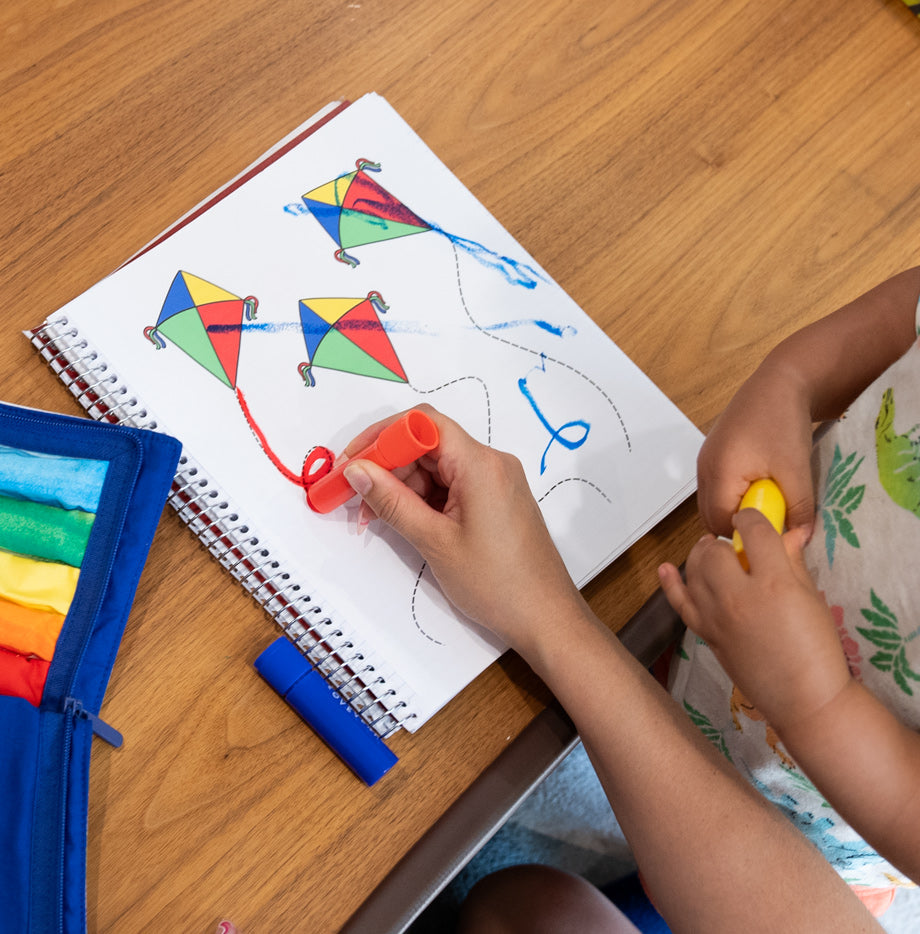 A child and an adult hand are seen interacting with a kite coloring activity in a spiral notebook on a wooden table. The child is coloring the strings of the kites while the adult holds a red crayon, assisting.