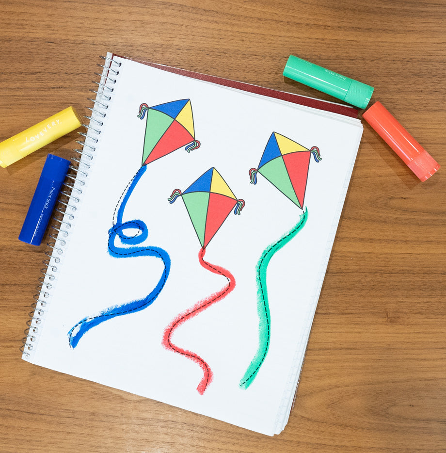 A completed activity in a spiral notebook lying on a wooden table, showing three kites with brightly colored strings in blue, red, and green crayon. The kites are filled with vibrant colors matching the crayons visible beside the notebook.