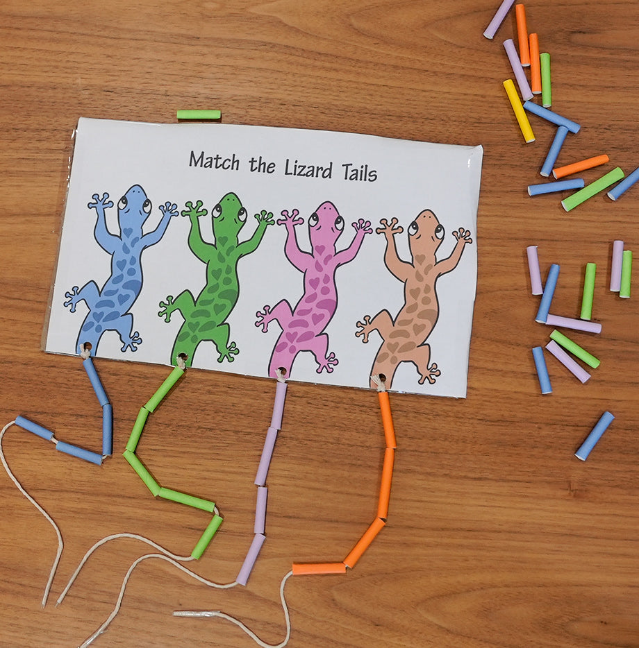 An activity sheet titled "Match the Lizard Tails" lies on a wooden table, partially completed with multicolored tubes and strings forming connections to simulate the tails of differently colored cartoon lizards.